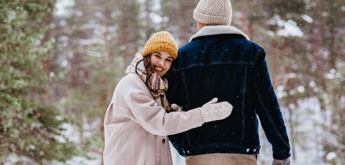 couple walking away in a snowy woods with woman looking back and smiling while hugging the man