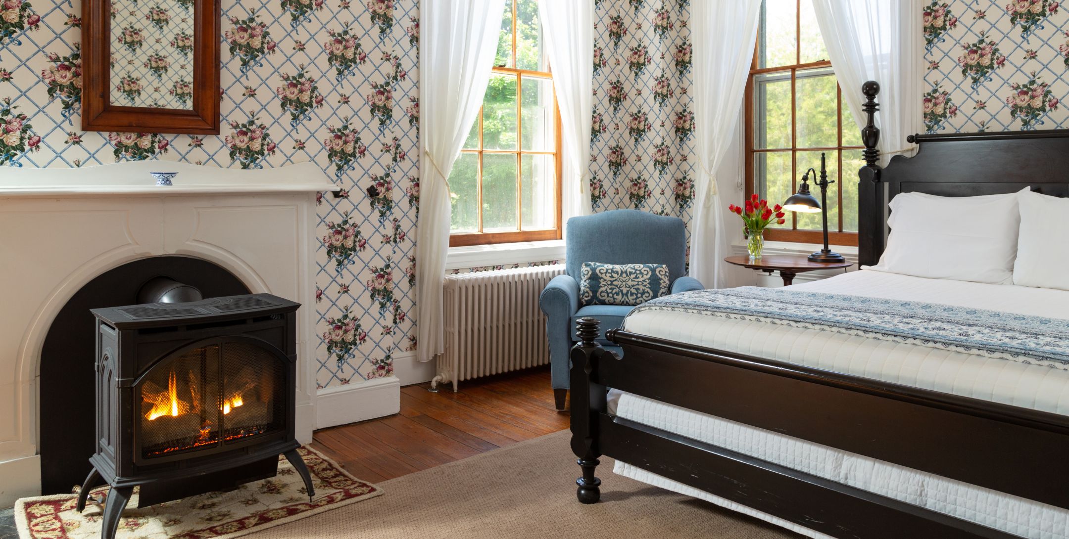 Wooden bed, hardwood floors, wallpapered walls, big window, fire stove, and comfy chair