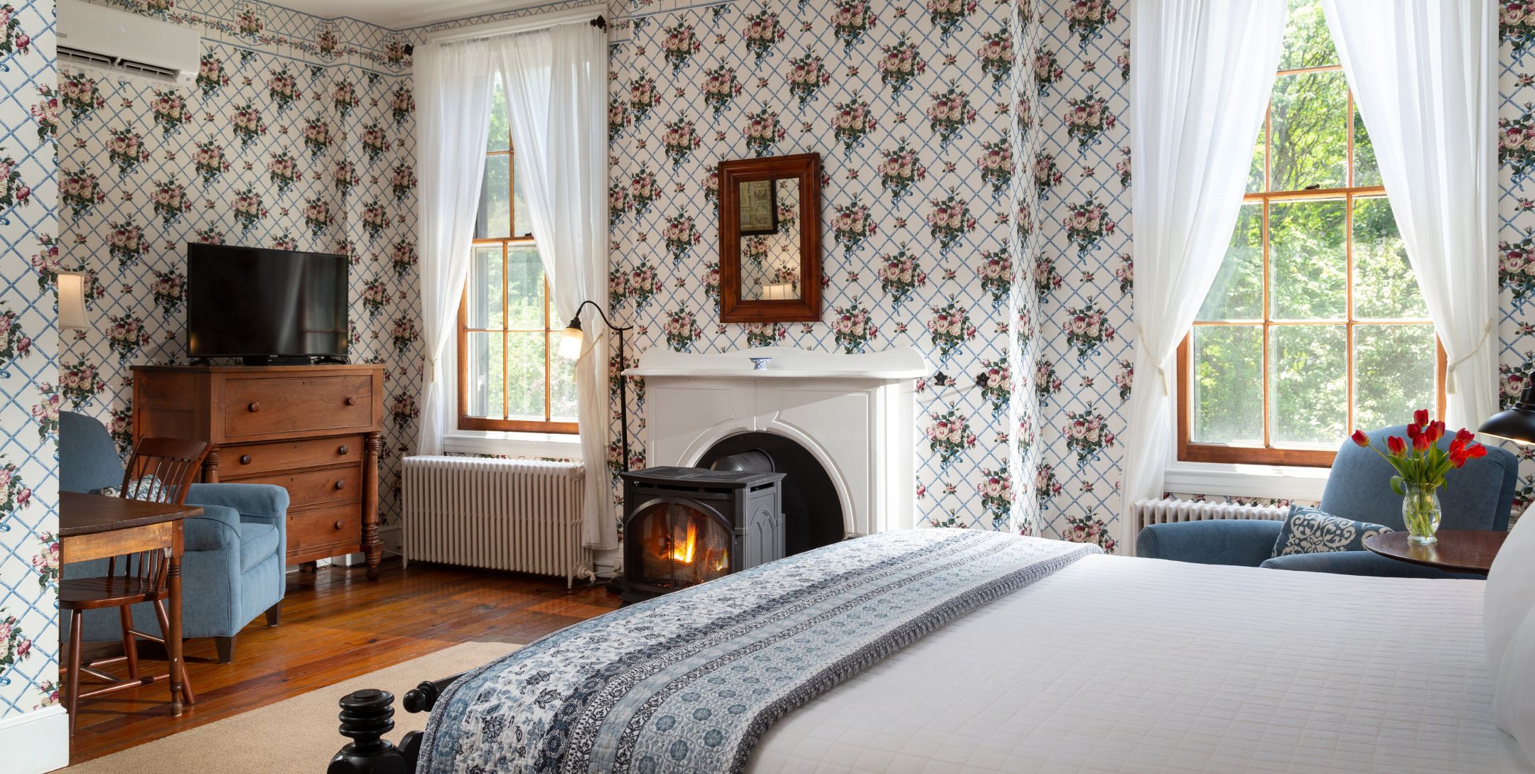 Spacious guest room with floral wallpaper, natural light, fire stove, table for two, TV and chair, and comfy bed