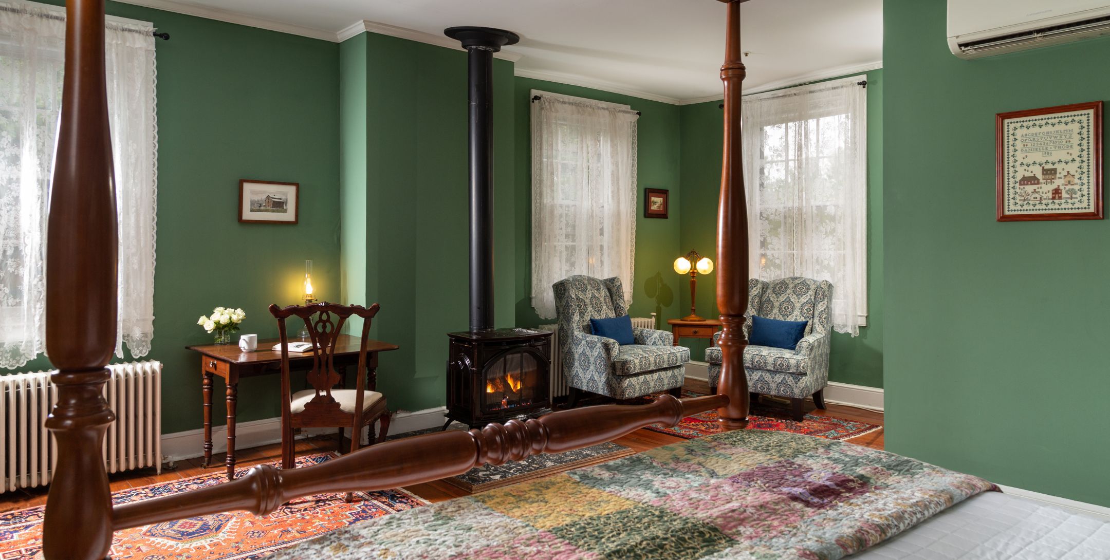 Guest room with deep green walls, hardwood floors, white lace curtains, two wing chairs and four poster bed