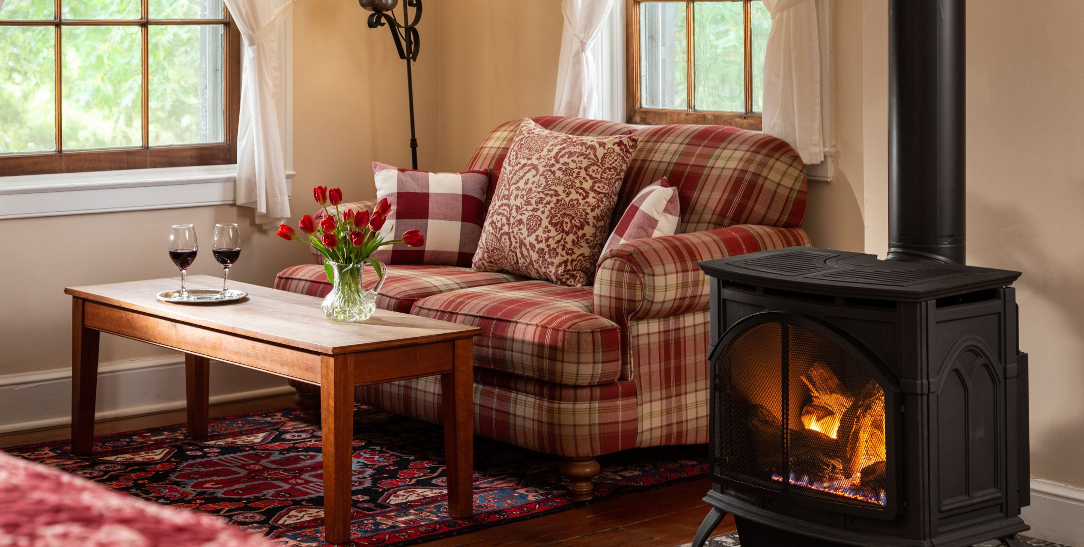 Red plaid loveseat in the corner flanked by two windows, with a coffee table topped with fresh flowers and two glasses of wine.