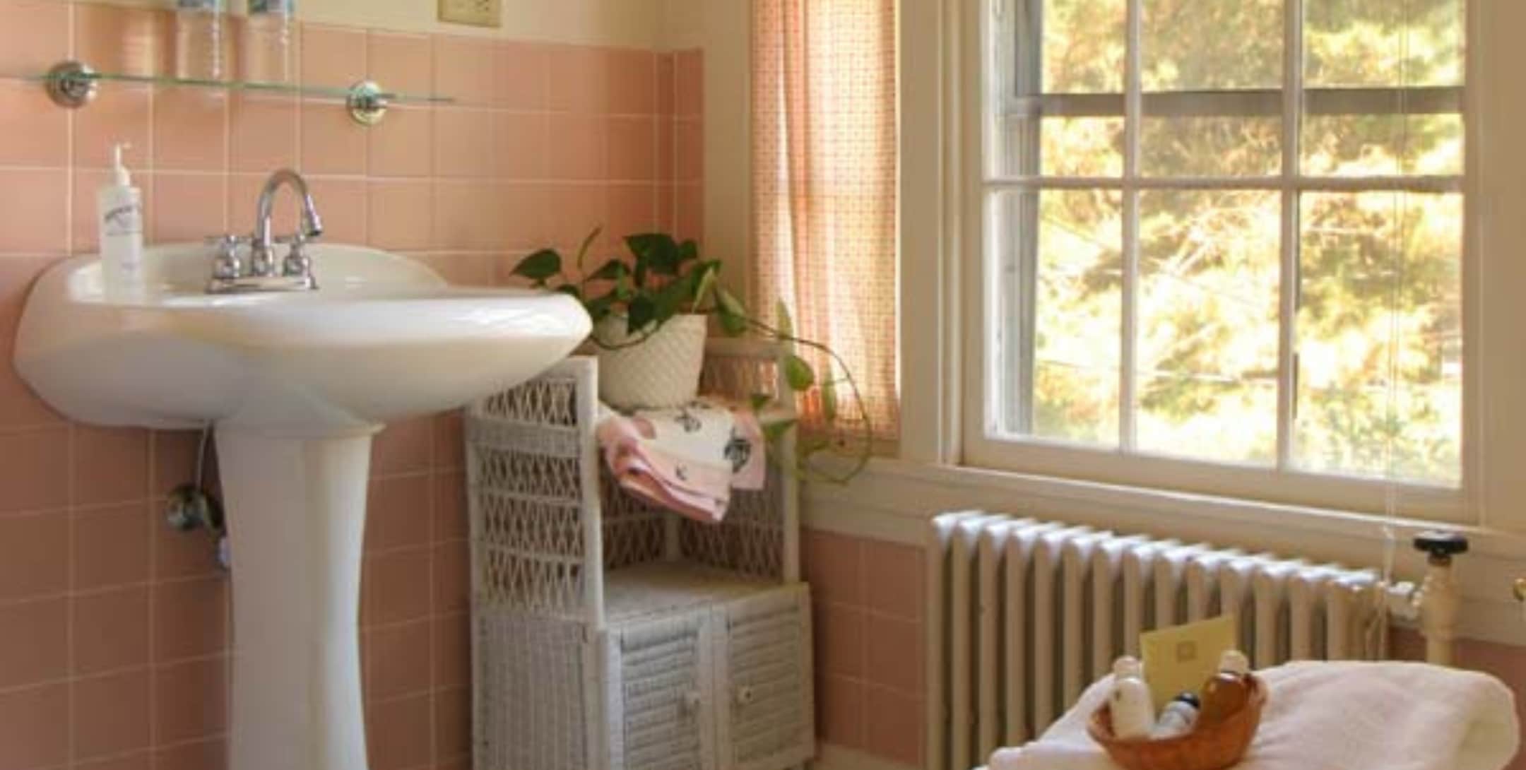 Poppy's bathroom with beige walls, pink tile, white pedestal sink and large window with sheer valance.