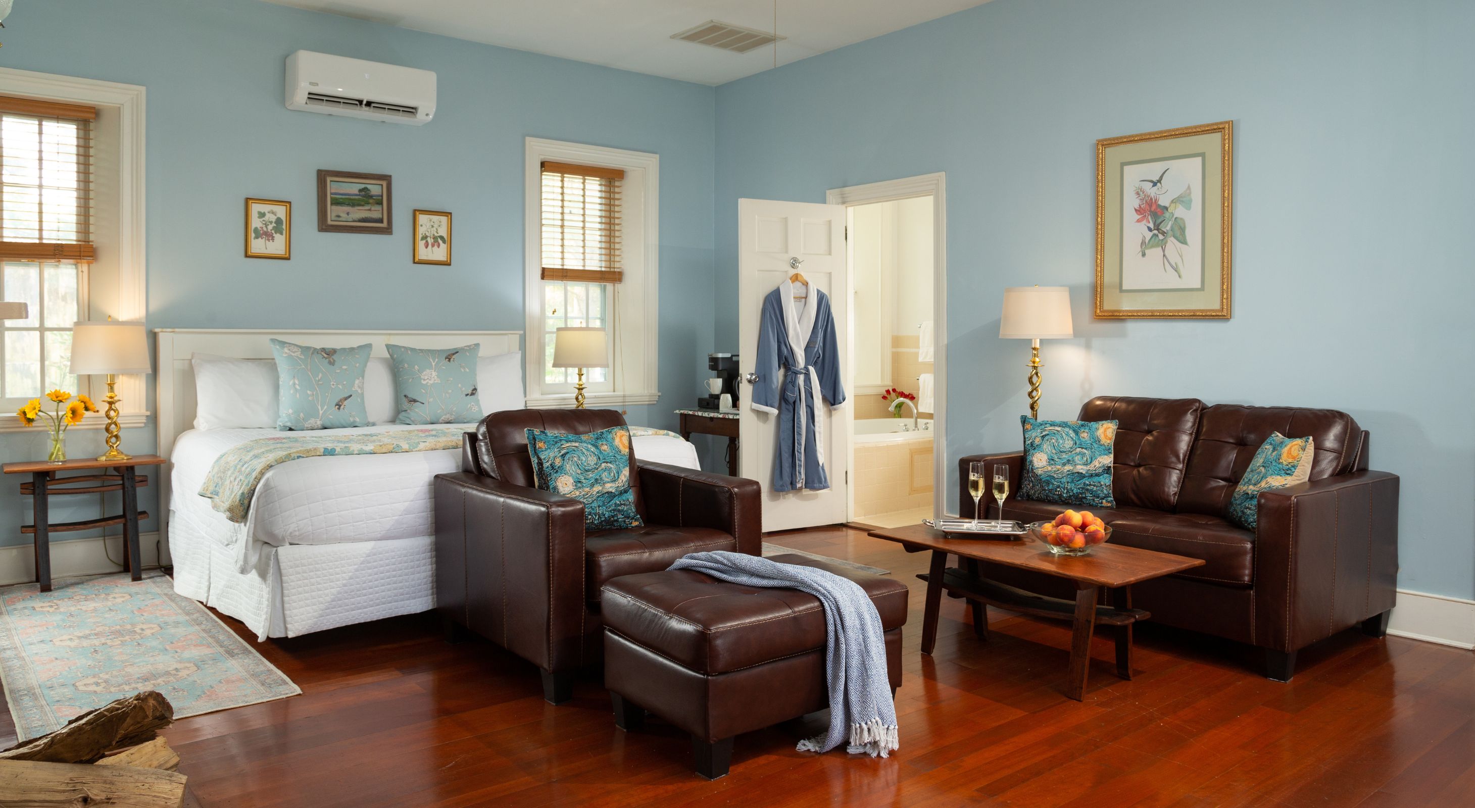 SLight blue Sunrise Room with off-white king bed, windows with blinds, wood floors, and leather sitting area.