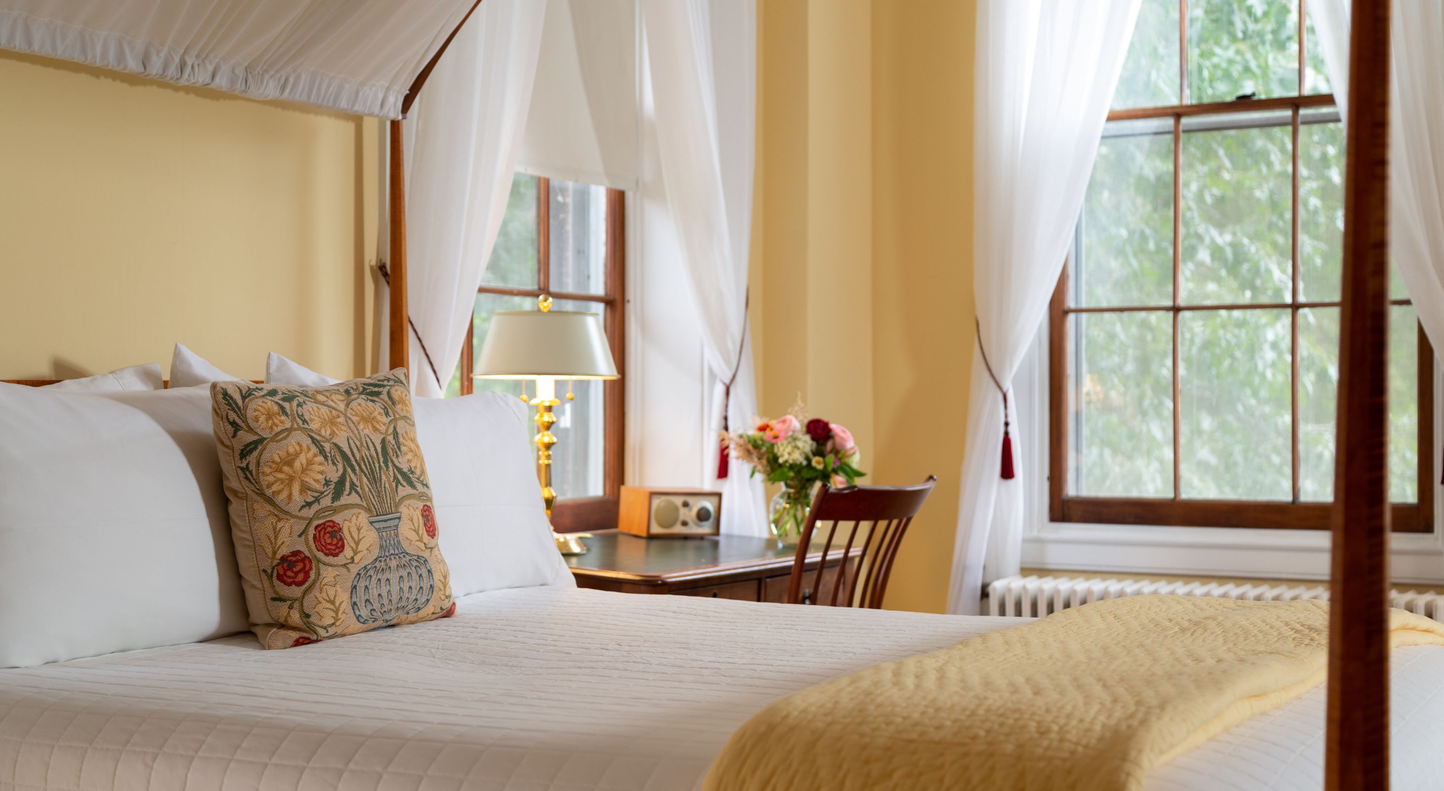 Close-up of canopy bed with white bedding and canopy, buttery yellow walls, and a desk near two windows with white shears