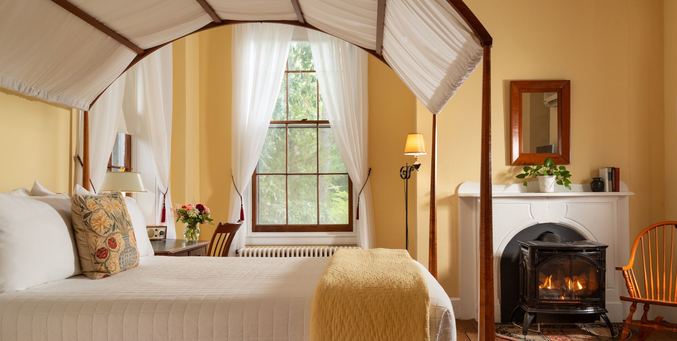 Close-up of canopy bed with white bedding and canopy, buttery yellow walls, and a desk near two windows with white shears