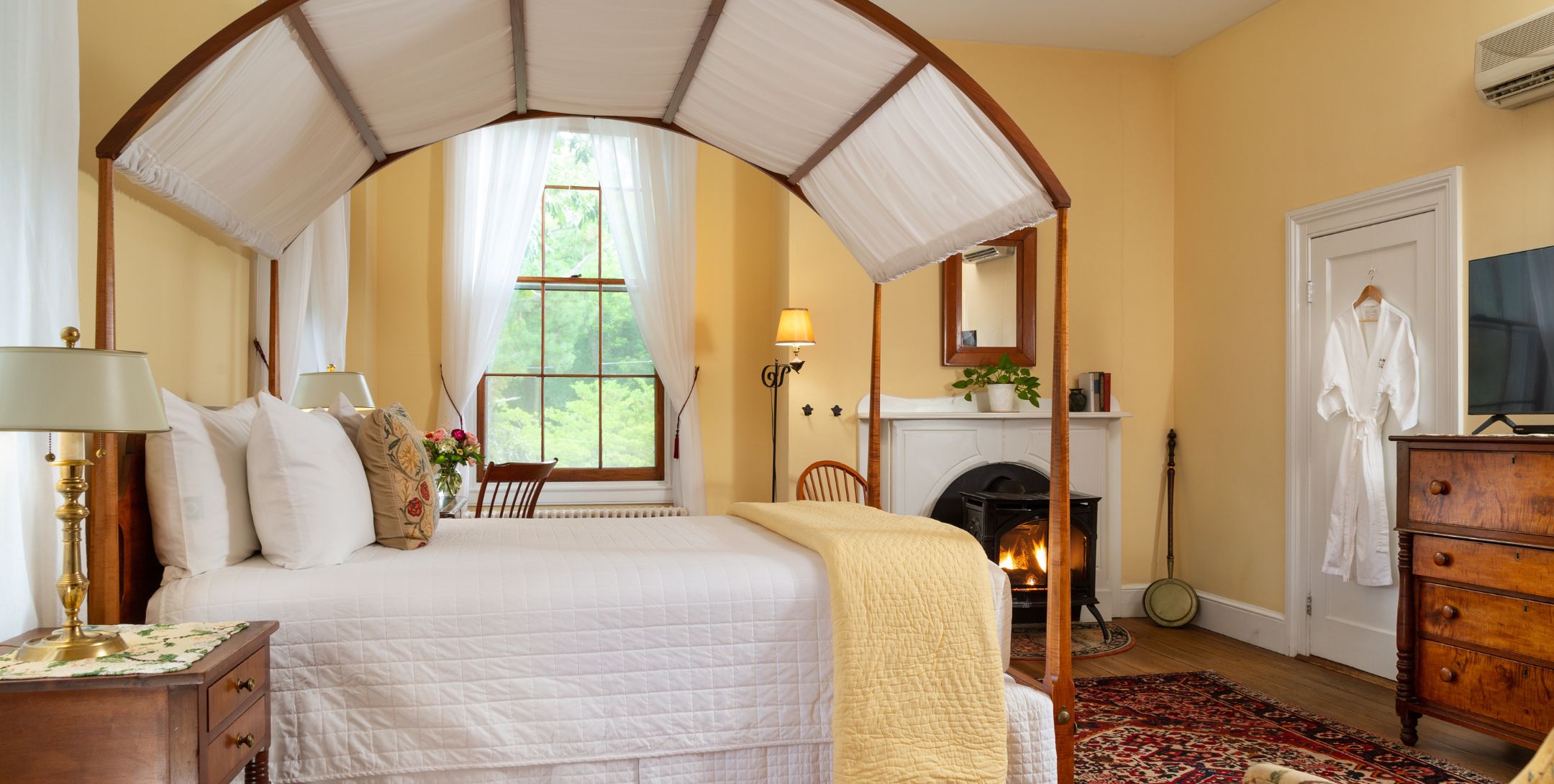 Buttery yellow guest room with natural light, fire stove, and arched canopy bed with white bedding and canopy