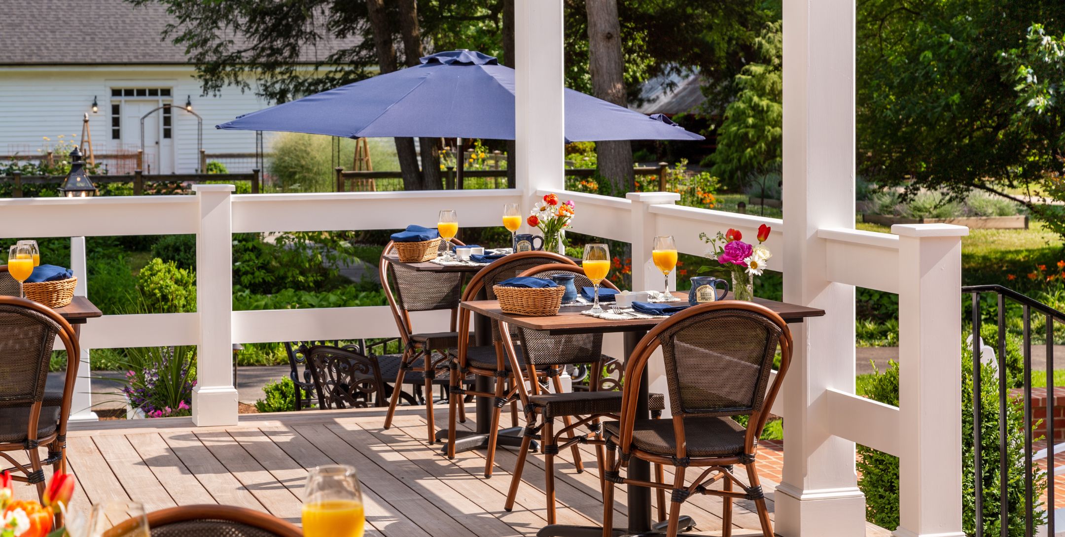 Outdoor dining deck with wicker tables for two set with orange juice in wine glasses, surrounded by lush landscaping