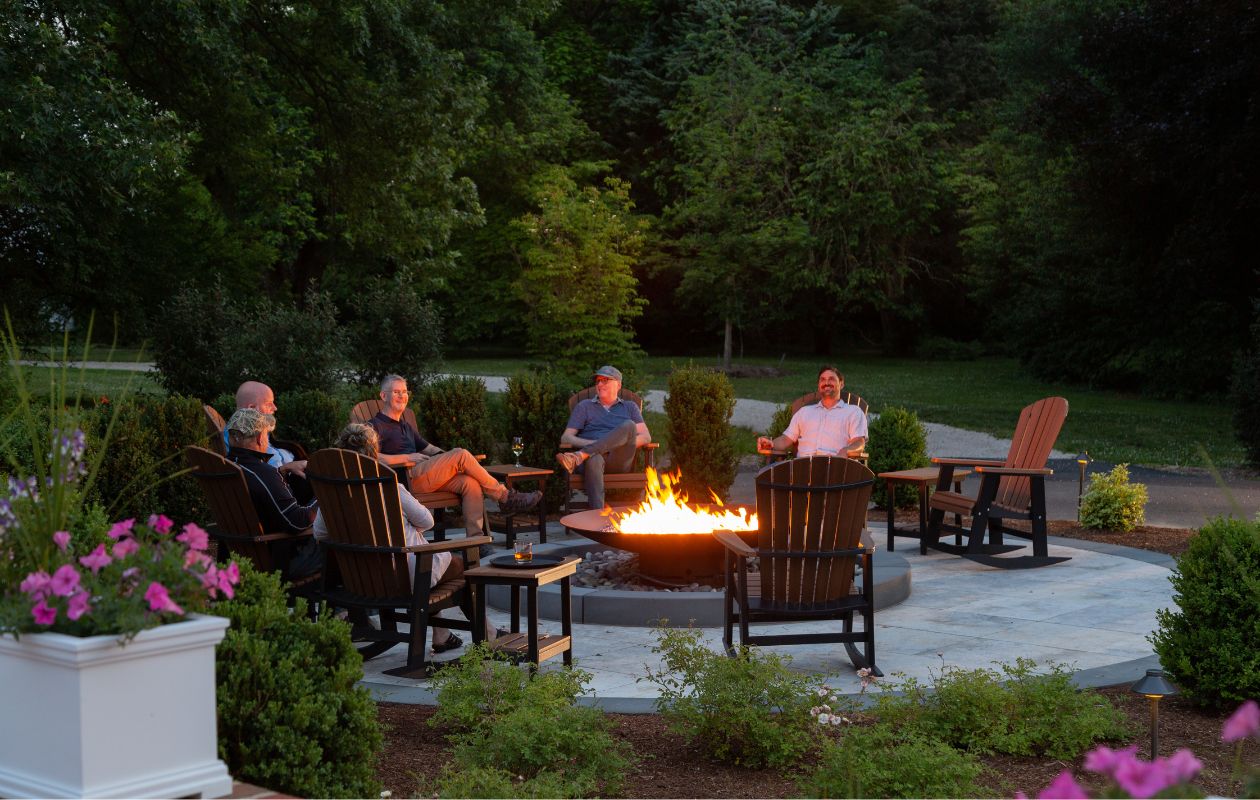 Group of people sitting in wooden Adirondack chairs around a fire pit on a round patio at dusk surrounded by lush landscaping