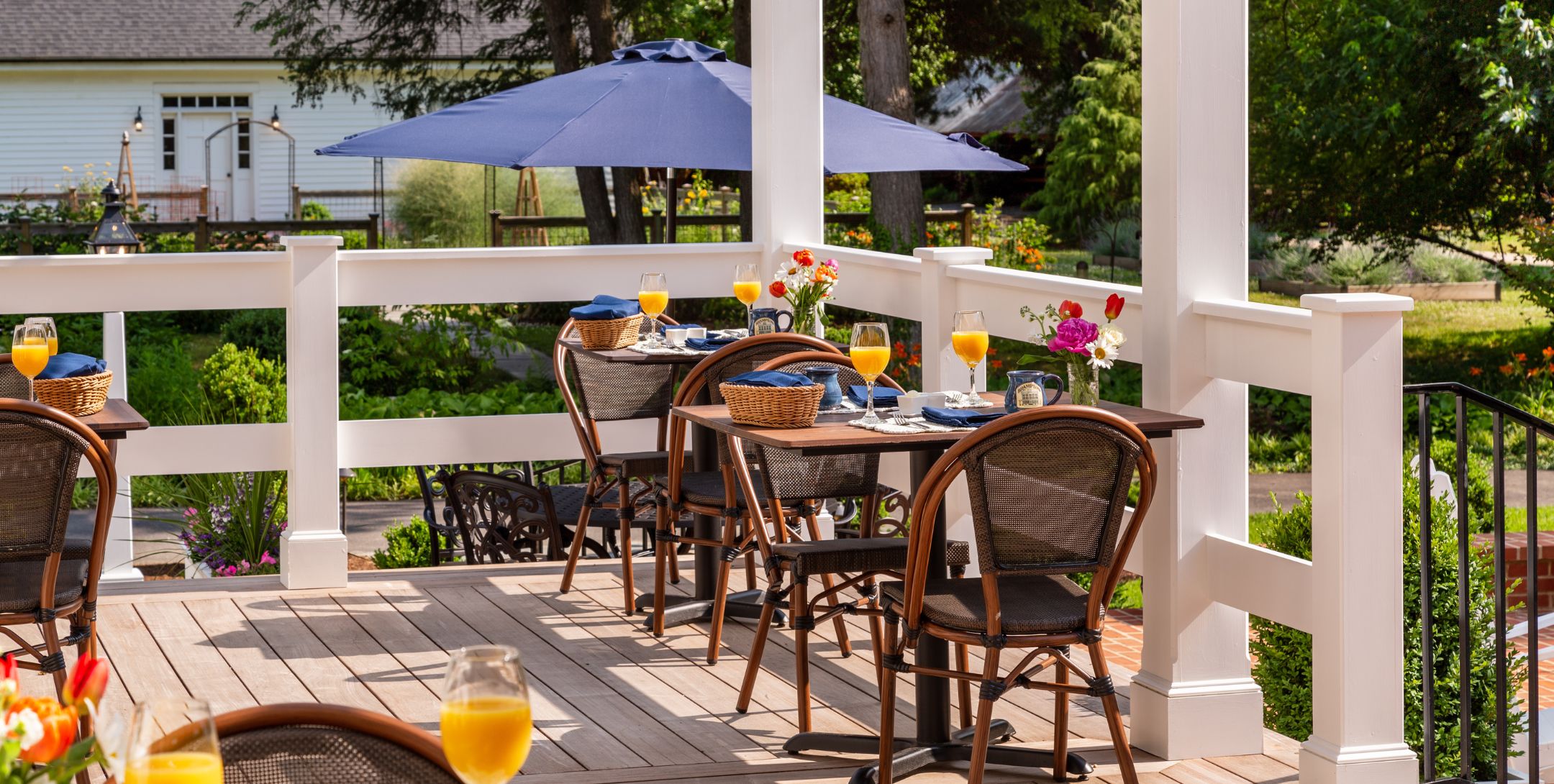 Outdoor dining deck with wicker tables for two set with orange juice in wine glasses, surrounded by lush landscaping