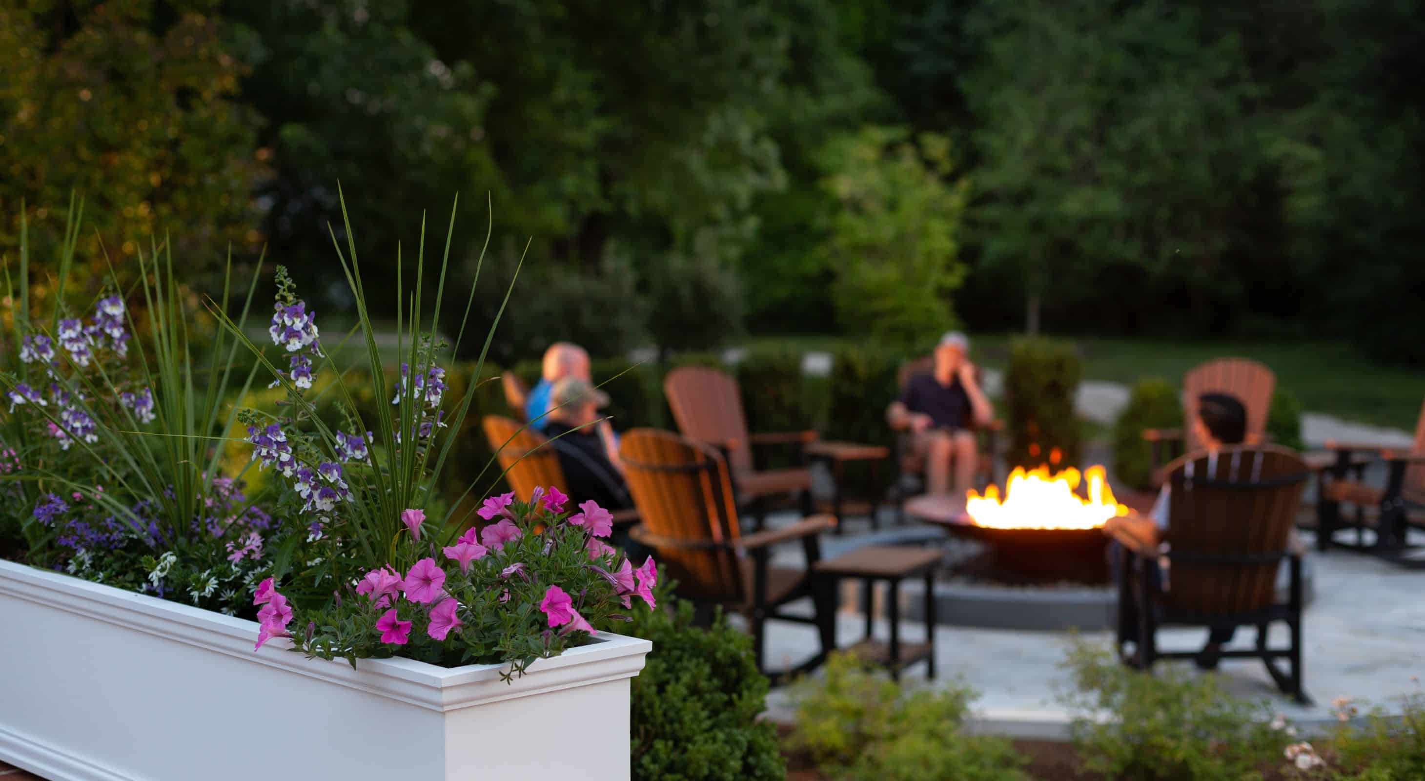 White flower box with pink flowers and group of people sitting in wooden Adirondack chairs around a fire pit in the background