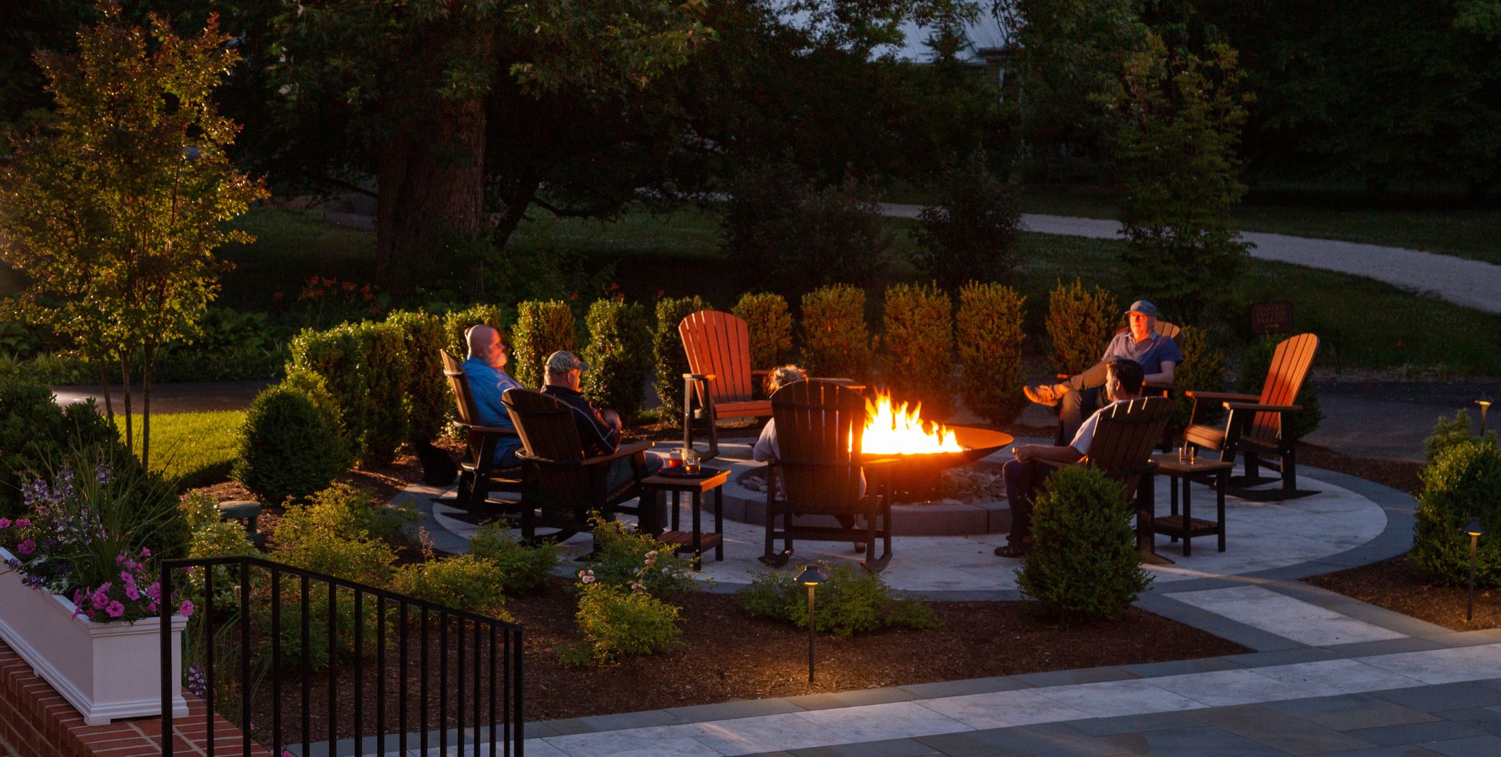 Group of people sitting in wooden Adirondack chairs around a roaring fire at dusk surrounded by lush green landscaping