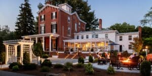 Exterior of multi-level inn at dusk with white portico, and guests by the fire.