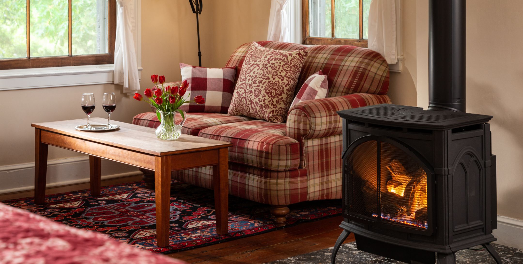 Red plaid loveseat in the corner flanked by two windows, with a coffee table topped with fresh flowers and two glasses of wine.