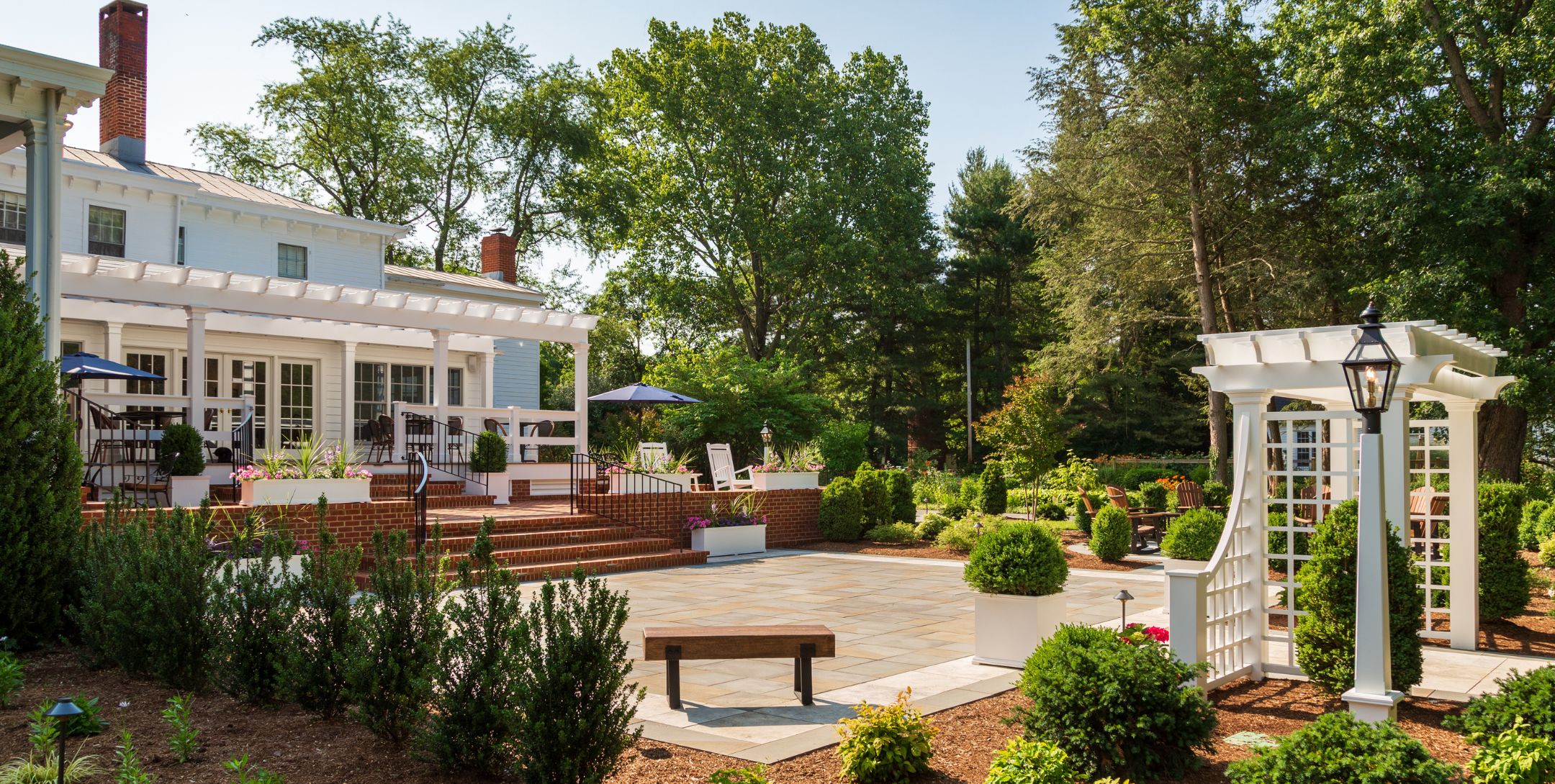 Rear exterior of inn with white portico, large stone patio, white trellis and lush green landscaping