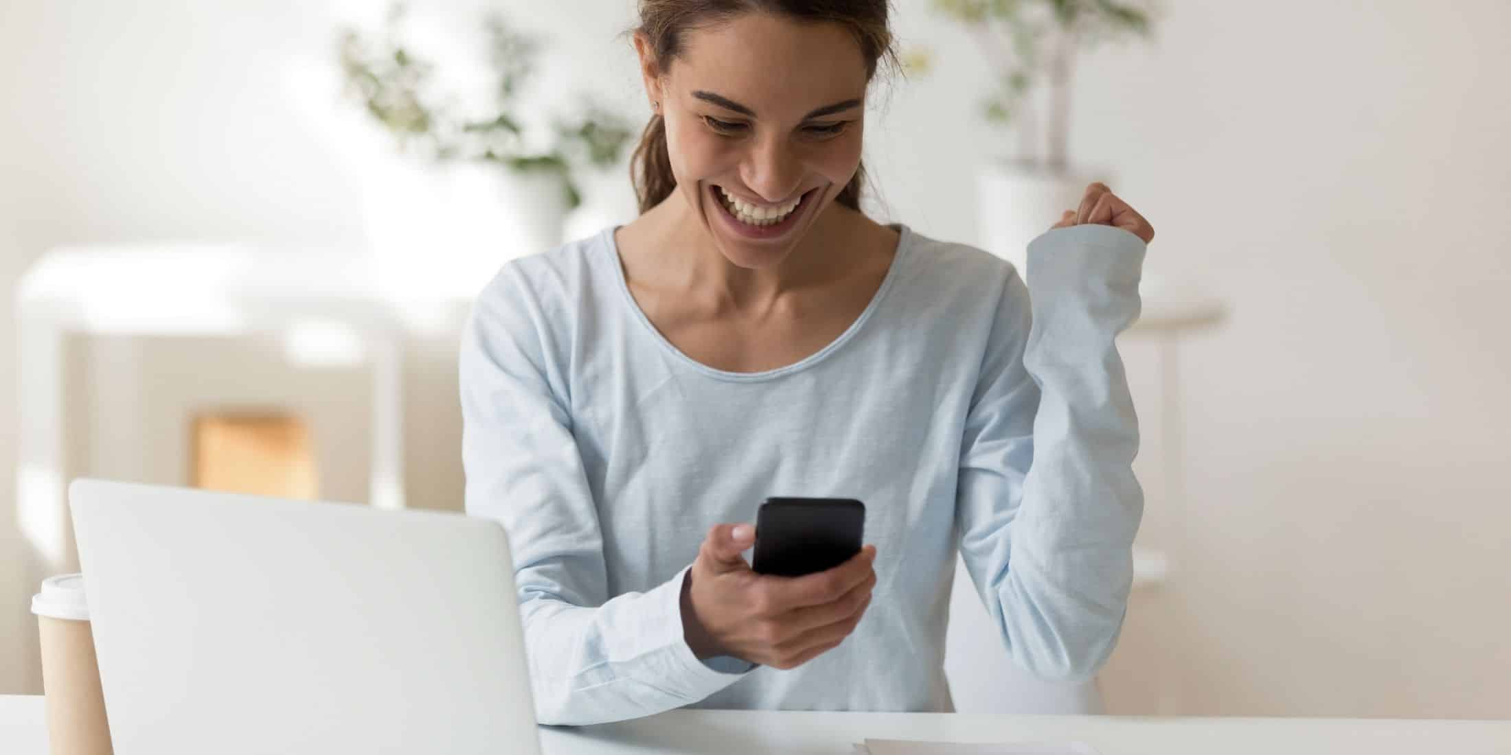 young woman smiling , looking down at mobile device she is holding in one hand. white computer open in front of woman and white walls behind her