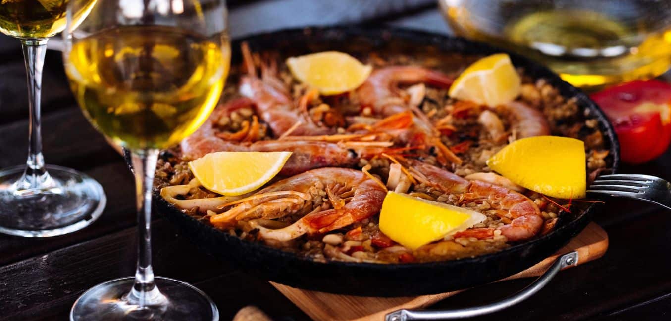 skillet with shrimp, rice, and lemon wedges next to two glasses of wine 