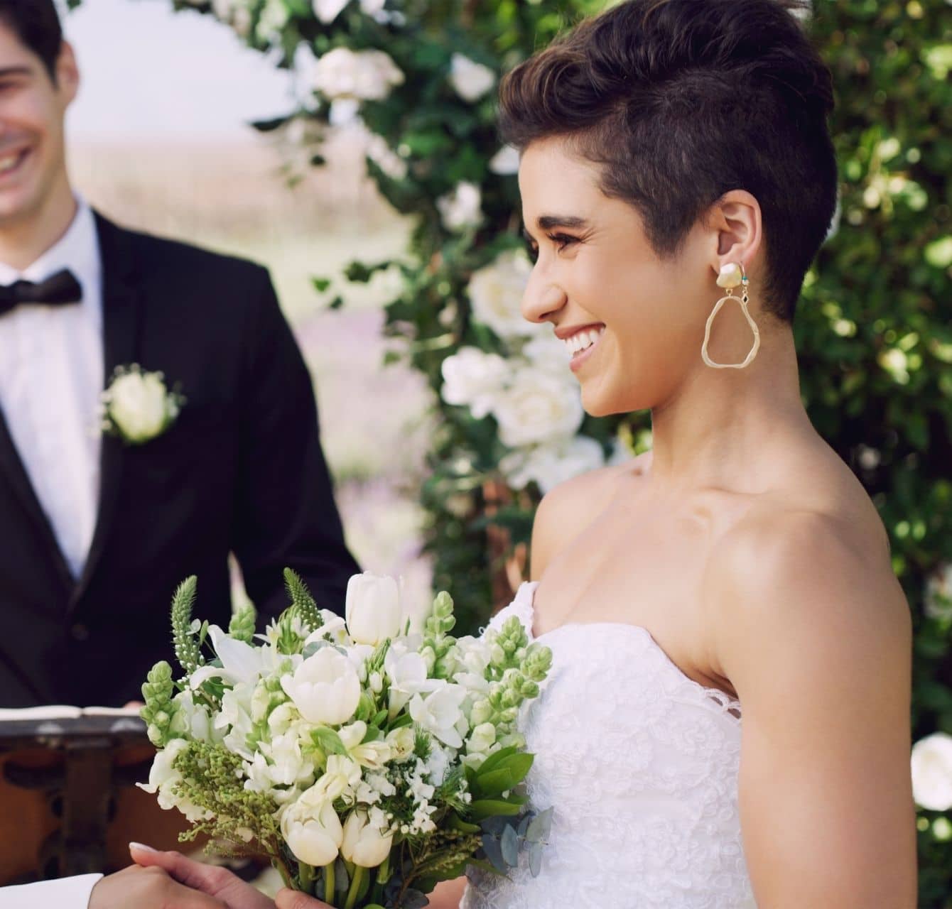 Two women in white bridal gowns facing each other. One is holding flowers. there is a man in tuxedo in the background as officiant