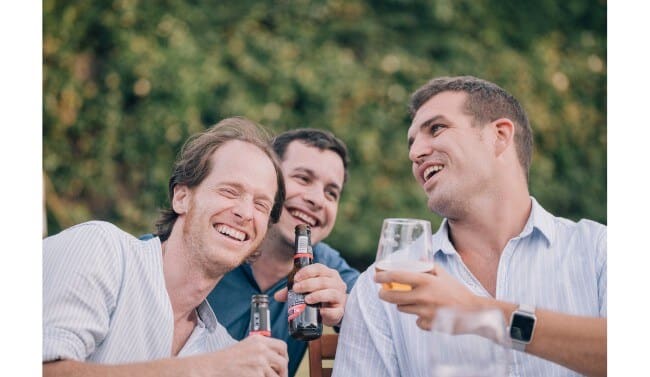 group of 3 smiling men holding beverages with woods behind